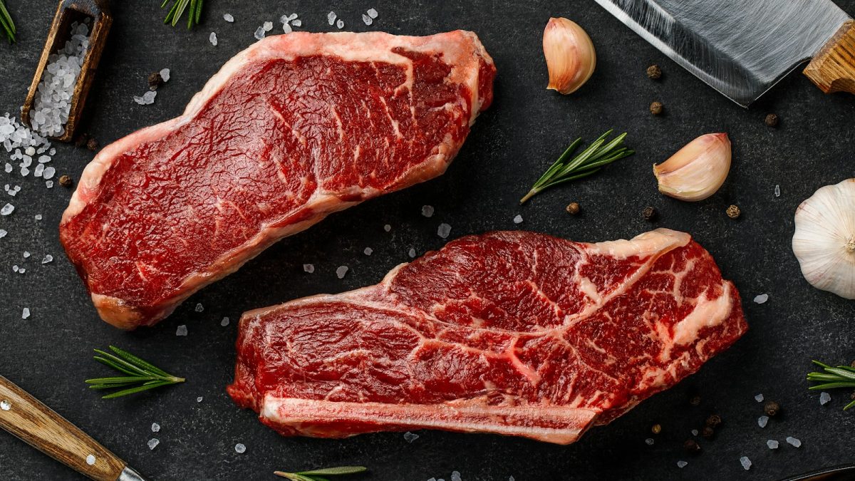 two steaks on a cutting board next to garlic, garlic, and a knife